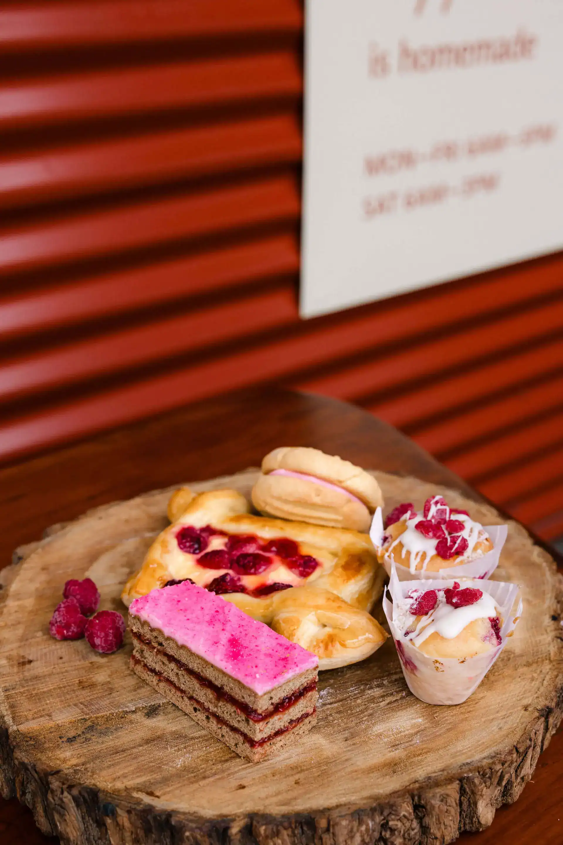 A wooden platter with assorted pastries, including a pink layered cake, fruit tart, cream-filled desserts, and raspberries, set on a wooden surface with a corrugated red background.