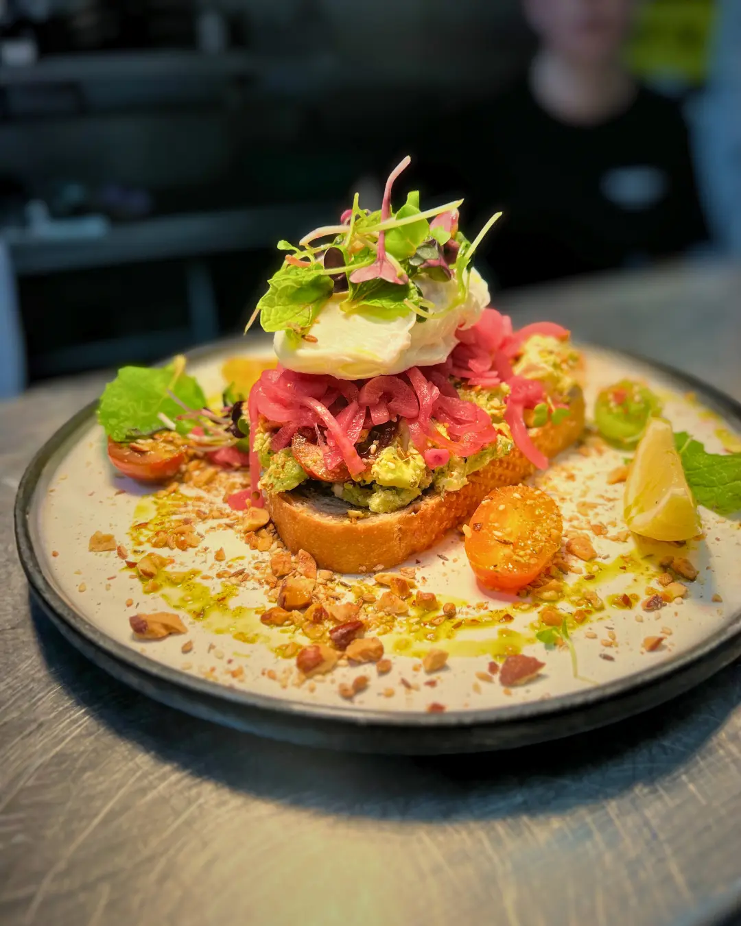 A plated dish featuring toast topped with avocado, pickled onions, a poached egg, microgreens, cherry tomatoes, chopped nuts, and garnished with a lemon wedge.