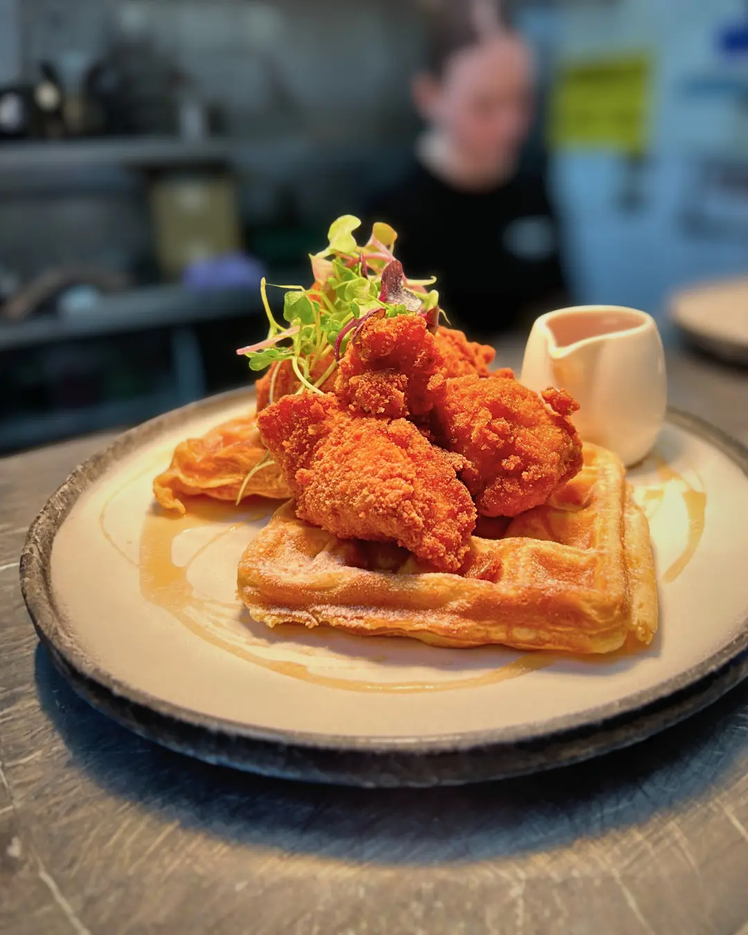 A plate of crispy fried chicken on waffles, topped with microgreens and served with a small pitcher of syrup.