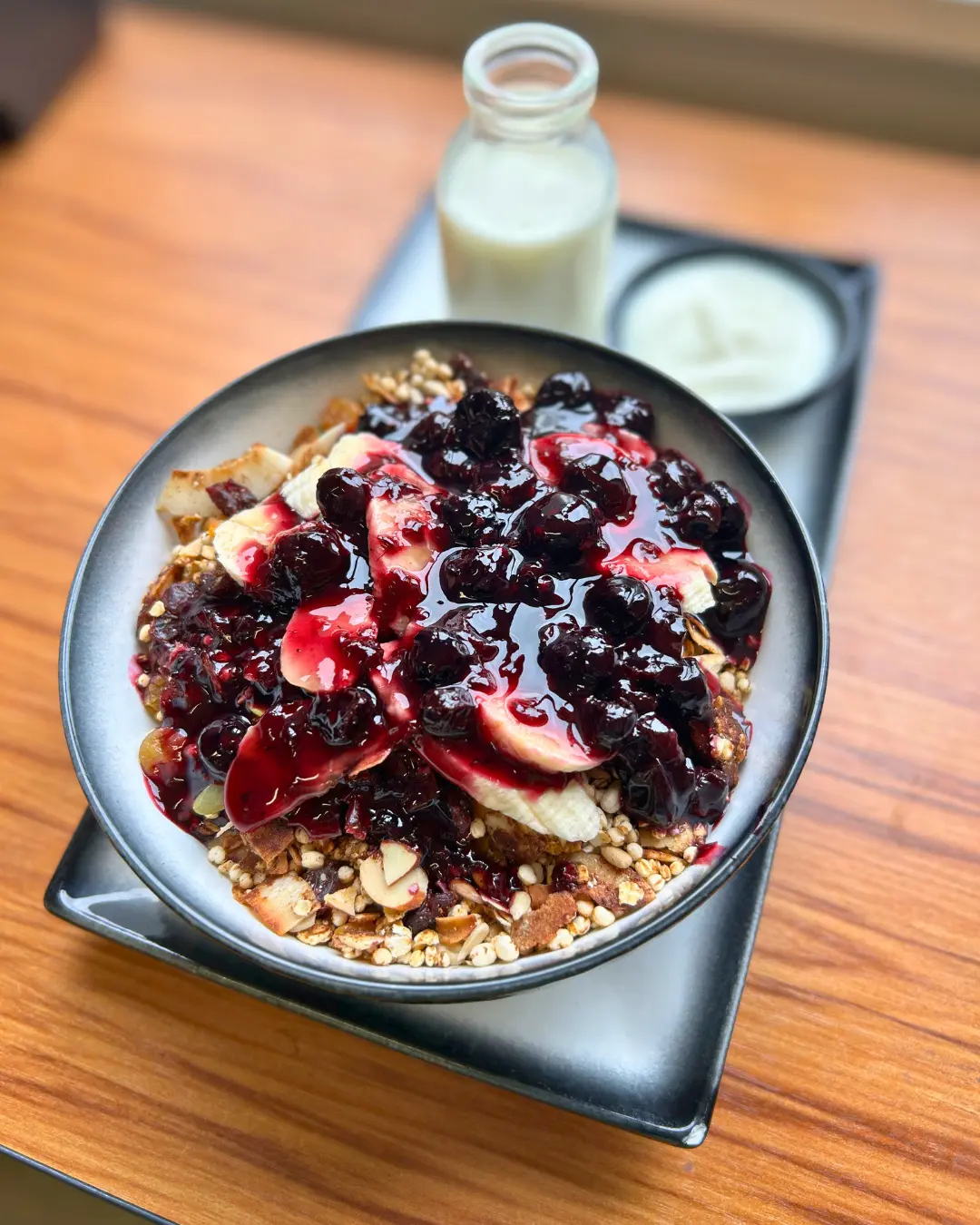 A bowl of granola topped with banana slices and berry compote, served with a small bottle of milk and a dish of yogurt on a wooden table.