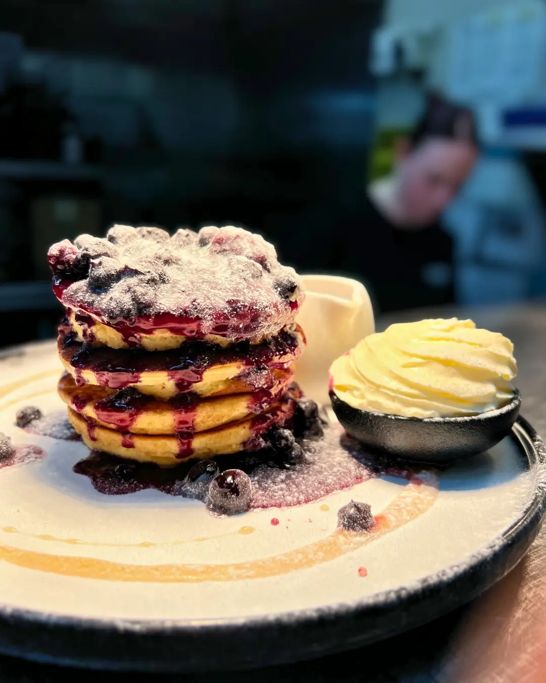 A stack of pancakes with blueberry sauce and powdered sugar, served with a small bowl of whipped cream on a plate; a blurred person in the background.