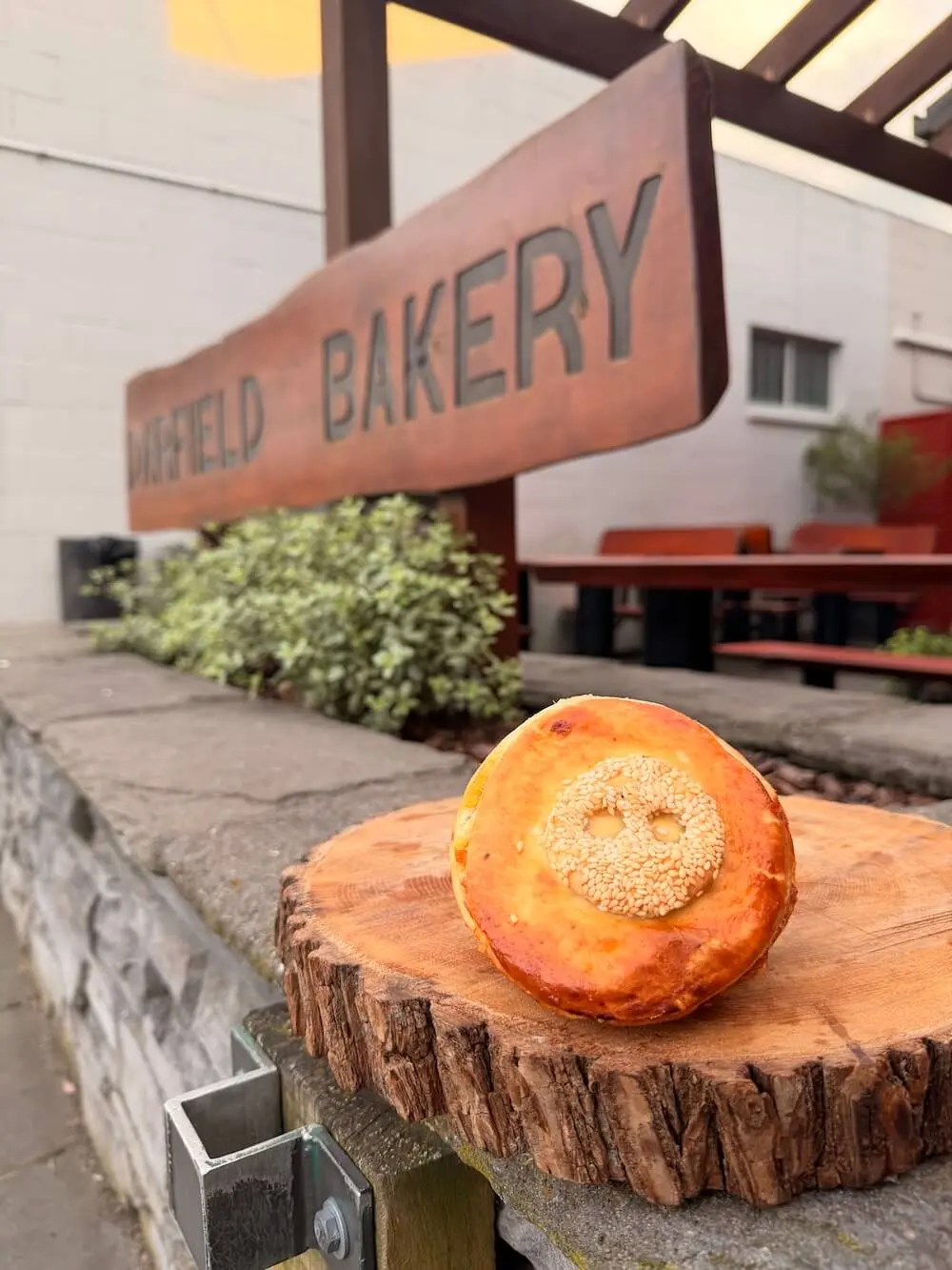 A round pastry with a sesame seed smiley face sits on a wooden board outside a bakery with a sign reading "BAKERY" in the background.