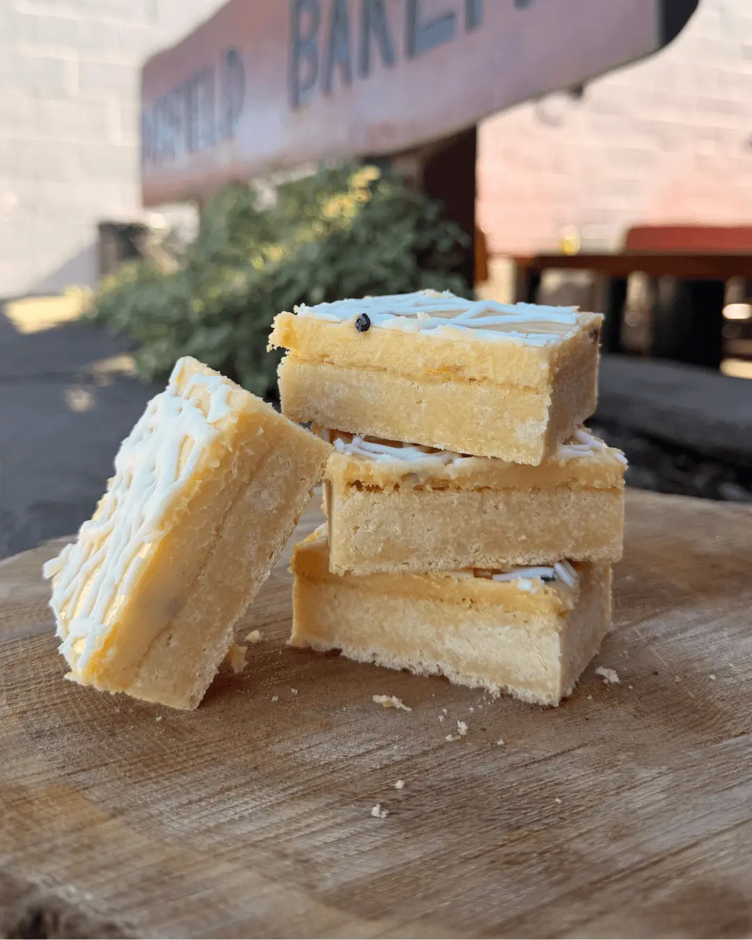 Four stacked passionfruit bars with white icing drizzle sit on a wooden surface outdoors, with a blurred bakery sign and plants in the background.
