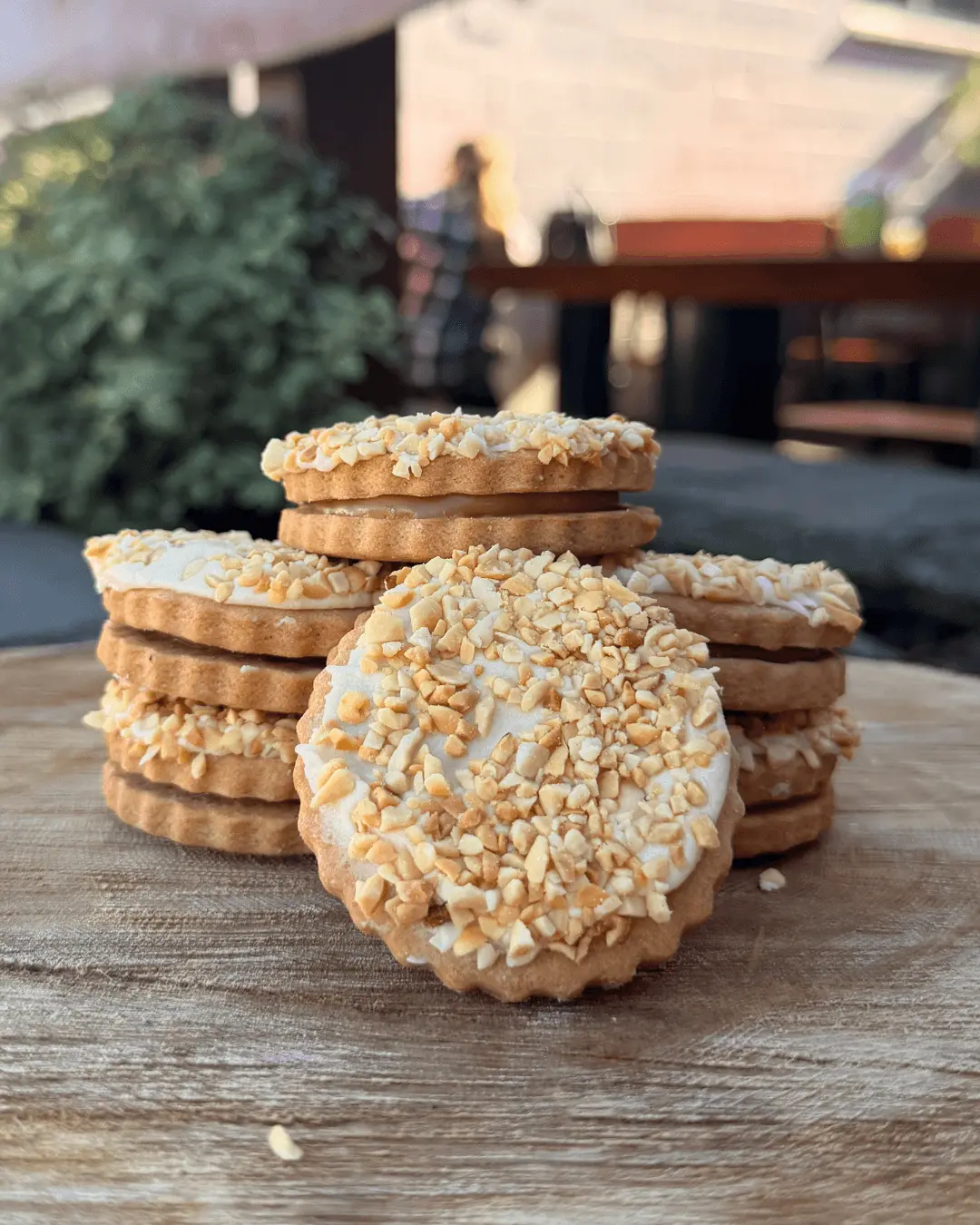A stack of round sandwich cookies topped with crushed nuts is arranged on a wooden surface outdoors.