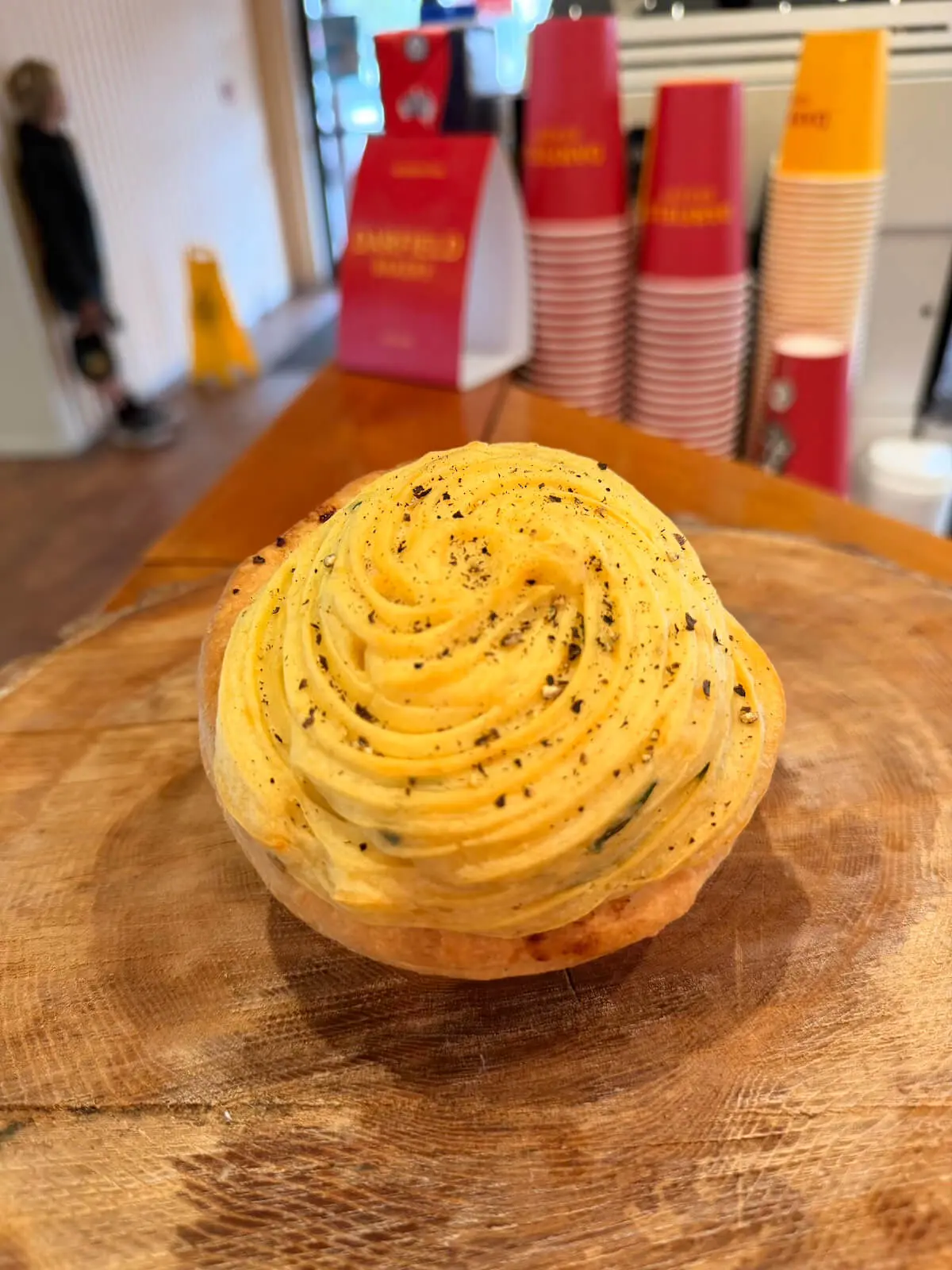 A savory pastry topped with piped mashed potatoes and black pepper sits on a wooden surface; stacked red and white cups are visible in the background.
