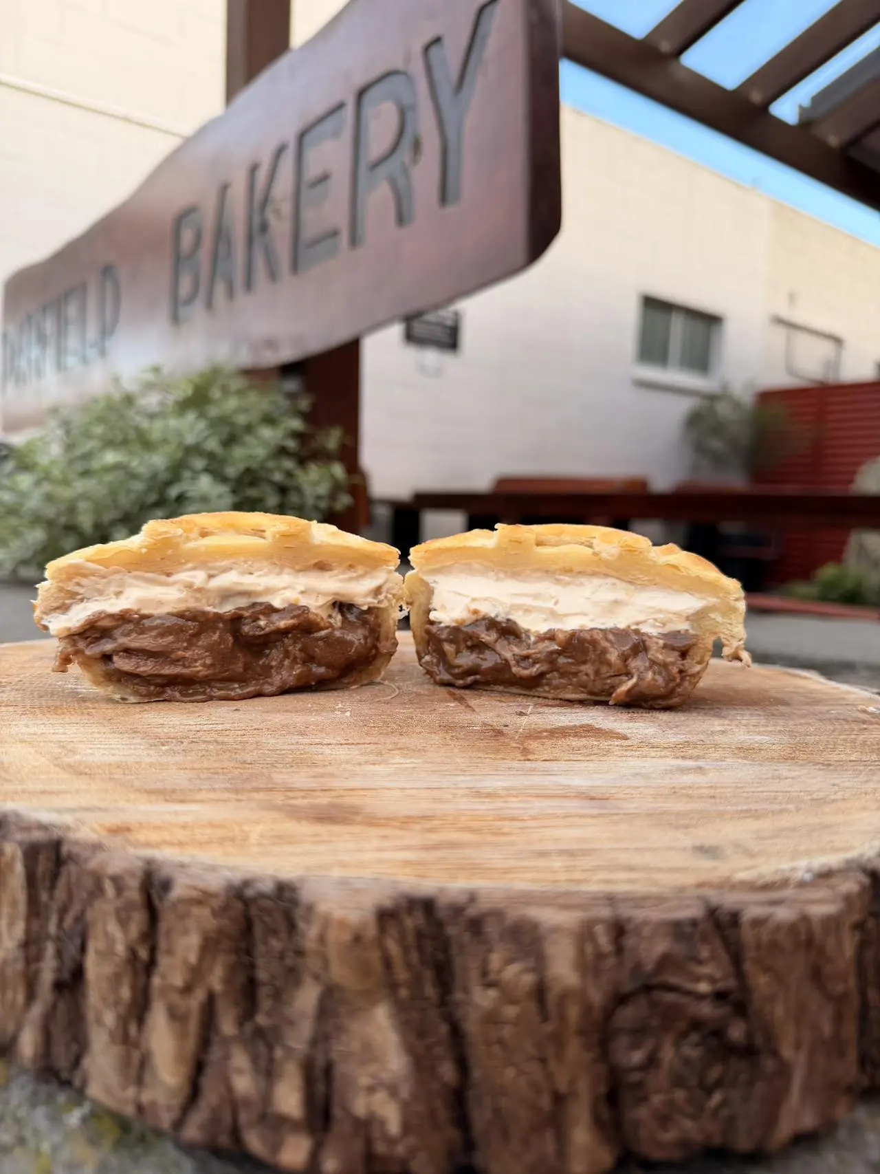 Two waffle pies with chocolate and cream filling are displayed on a wooden slab outdoors, with a bakery sign visible in the background.