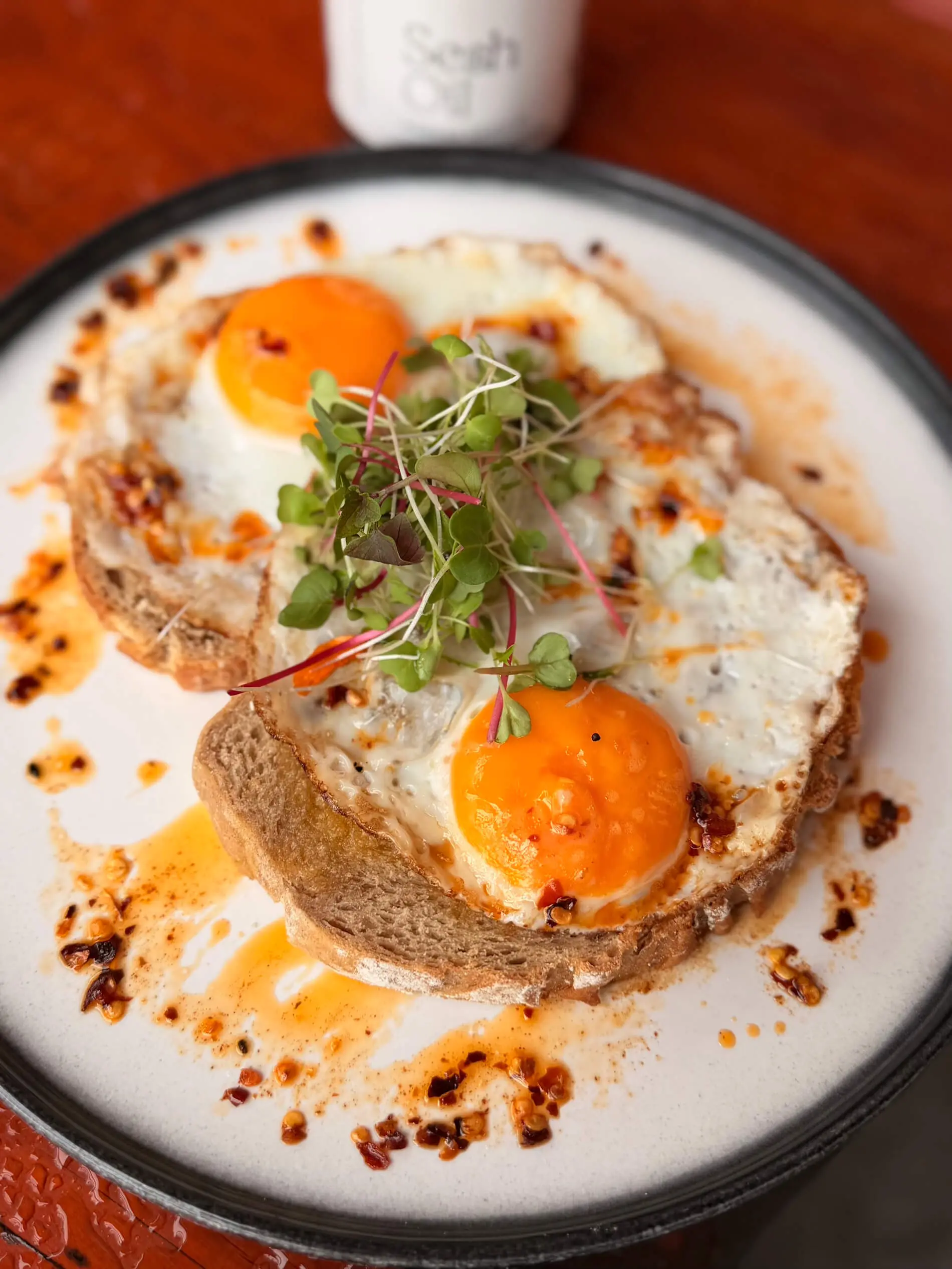 Two sunny side up eggs on toasted bread, topped with microgreens and chili oil, served on a white plate.
