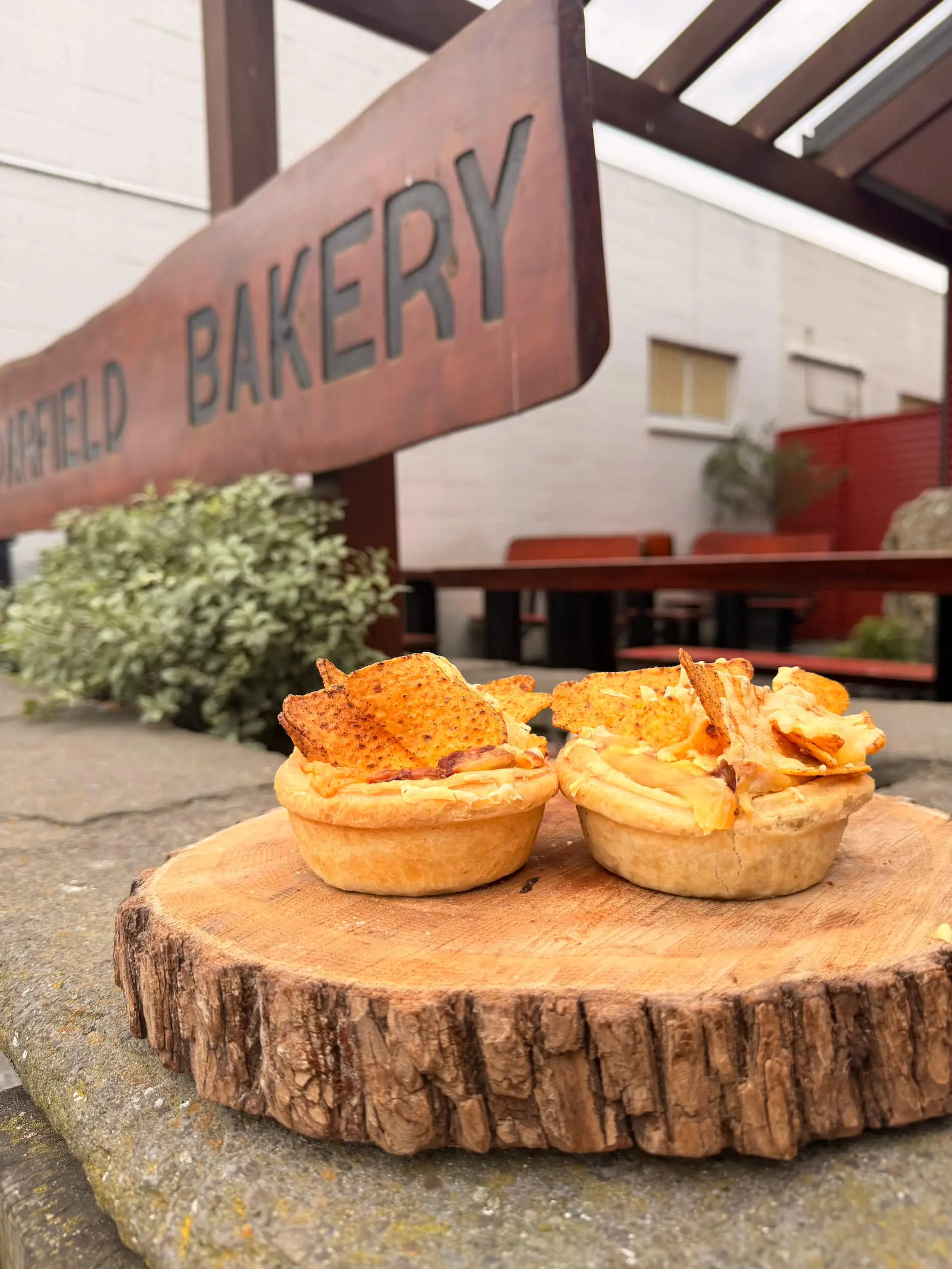 Two savory pies topped with chips are displayed on a wooden board outside a bakery with a rustic sign in the background.