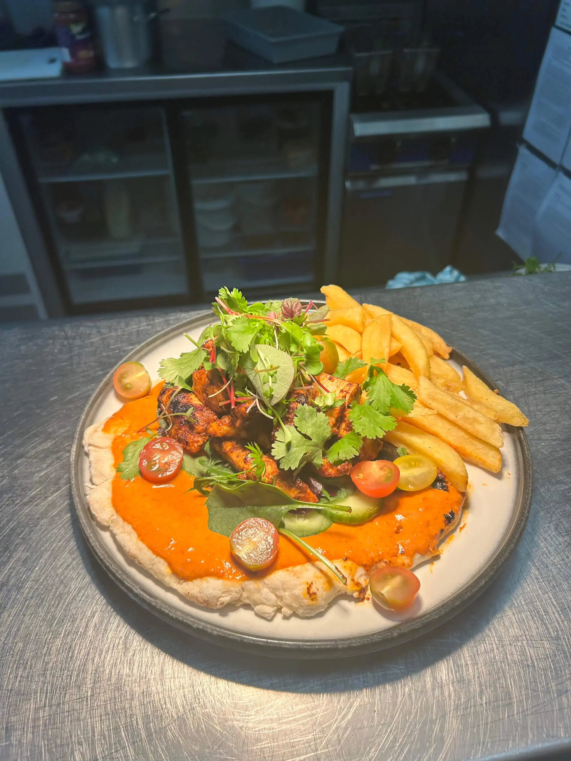 A plate with seasoned grilled chicken on flatbread with red sauce, cherry tomatoes, greens, and a side of fries, served on a stainless steel counter in a kitchen.