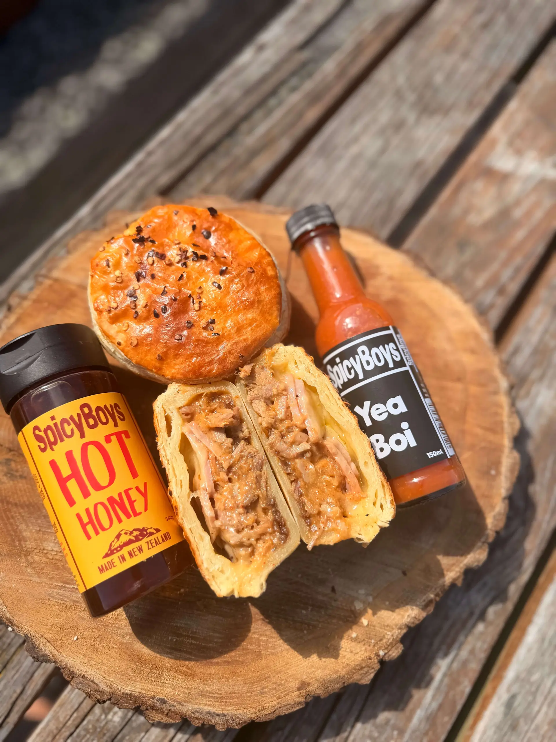 Round meat pie cut in half on a wooden board, with Spicy Boys Hot Honey and Yea Boi hot sauce bottles placed beside it.